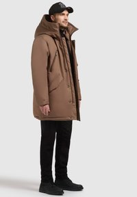 Braune isolierte Parka mit hohem Kragen, mehreren Reißverschlusstaschen und Knopfverschlüssen, kombiniert mit schwarzen Hosen und schwarzen Schuhen.