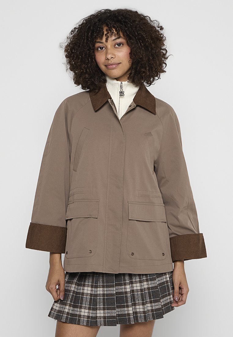 Jonge vrouw met krullend haar die een taupe jas met een bruin kraag en manchetten draagt over een witte rits-top en een geruite rok.