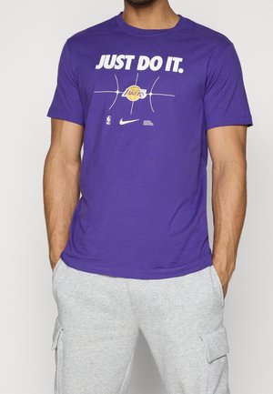 Homme portant un t-shirt violet des NBA Los Angeles Lakers avec le texte "JUST DO IT" et un pantalon de survêtement gris avec des poches, mains dans les poches.