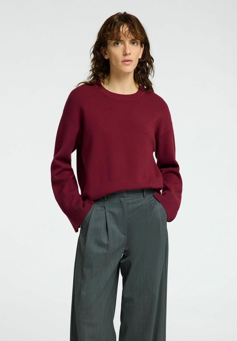 Bordeauxfarbener Pullover mit Rundhalsausschnitt und weit geschnittenen Ärmeln, kombiniert mit grauen, gefalteten Hosen, beide aus weichen Materialien und maßgeschneiderter Passform.