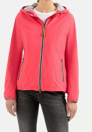 Femme portant une veste à capuche zippée corail vif par-dessus une chemise blanche et un jean noir, debout devant un fond uni.