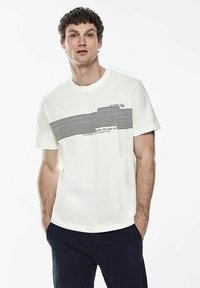 Bílý bavlněný tričko s vodorovnými černými pruhy a textem. Krátké rukávy, kulatý výstřih, volný střih. Modelka má na sobě námořnické kalhoty.