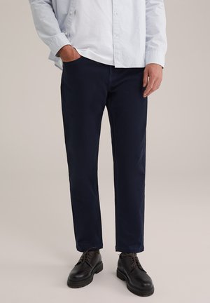 Marineblaue, gerade geschnittene Hose aus weichem Stoff, kombiniert mit schwarzen Schnürschuhen. Ein hellblaues Hemd wird über einem weißen T-Shirt getragen.