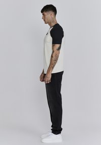 Camisa de manga corta para hombre con cuerpo crema y mangas raglán negras, combinada con jeans negros y zapatillas blancas. Presenta tatuajes visibles en los brazos.