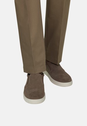 Bruine suède instapschoenen gecombineerd met kaki broek tegen een witte achtergrond, met zicht op onderbenen en voeten.