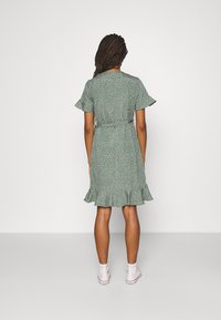 Vestido verde com bolinhas brancas, mangas curtas com babados, cinto na cintura e saia evasê. Usado com ténis brancos.