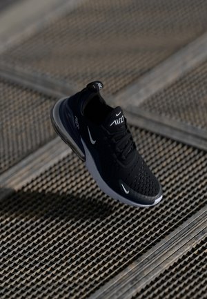 Basket Nike Air Max 270 noire avec logos swoosh blancs, flottant au-dessus d'un fond en grille métallique texturée.
