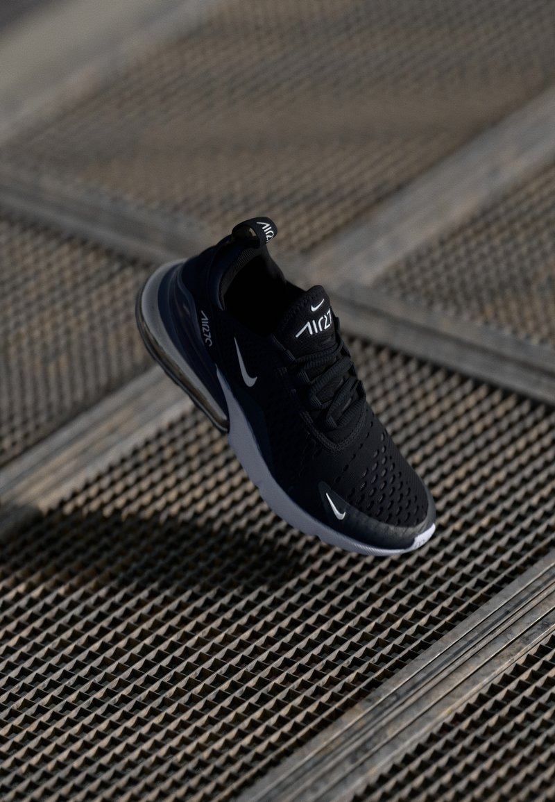 Basket Nike Air Max 270 noire avec logos swoosh blancs, flottant au-dessus d'un fond en grille métallique texturée.