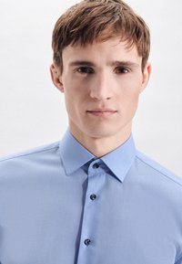 Camicia azzurro chiaro con colletto, maniche corte e tre bottoni scuri lungo la parte anteriore. Tessuto liscio e design aderente.