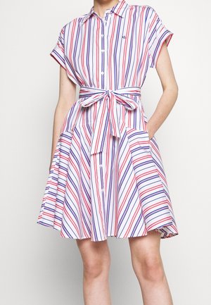 Femme portant une robe courte à manches courtes, longueur genou, avec des rayures verticales rouges et bleues et une ceinture nouée à la taille, les mains dans les poches.