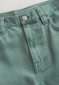 Gros plan sur la ceinture d'un jean en denim vert taille droite avec un bouton en métal et des passants pour ceinture.
