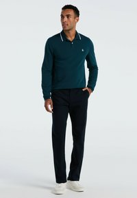 Teal langwerpige polo shirt met witte kraagaccenten, gecombineerd met donkere broeken en witte schoenen. Eenvoudig ontwerp, slim fit en een gladde textuur.