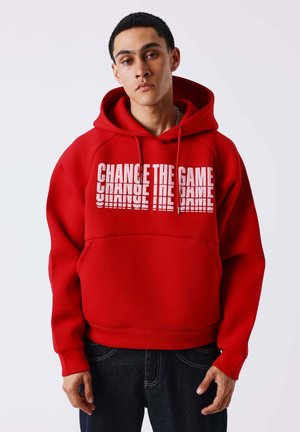 Joven con sudadera roja con texto "CHANGE THE GAME" en blanco, de pie frente a un fondo claro y liso.