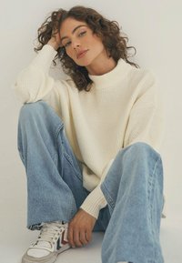 Cremfarbener, gerippter Strickpullover mit hohem Kragen, kombiniert mit hellblauen, weit geschnittenen Jeans und weißen Sneakers mit roten und blauen Akzenten.