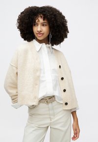 Beige pletený cardigan s ribbovým vzorem, s černými knoflíky. Nošený přes bílou košili s límečkem a v kombinaci s béžovými kalhotami.