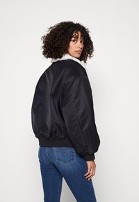 Czarna kurtka bomberka z kołnierzem z sztucznego futra, elastycznymi mankietami i dołem. Model nosi ją z dopasowanymi niebieskimi dżinsami. Gładka, błyszcząca tekstura.