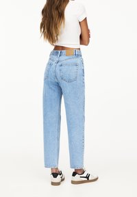 Femme portant un jean taille haute bleu clair, un t-shirt court blanc et des baskets blanches avec des détails noirs, debout les bras croisés.