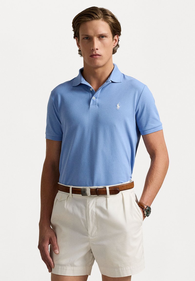 Cămașă polo albastră deschis, confecționată din bumbac, având un guler clasic, mâneci scurte și logo brodat. Asortată cu pantaloni scurți bej și o curea maro.
