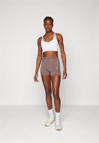 Reggiseno sportivo bianco con spalline incrociate, abbinato a pantaloni corti marroni a vita alta con cuciture a contrasto e un logo. Sneakers bianche e calze.
