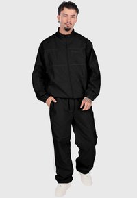 Melna sporta treniņtērps ar rāvējslēdzēja jaku un saskaņotām biksēm. Iespējas ietver elastīgas piedurknes un baltas sānu svītras uz biksēm. Gluda tekstūra.