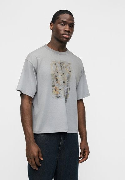 Νέος άνδρας που φοράει μια φαρδιά γκρι μπλούζα με αφηρημένο ανθισμένο σχέδιο και κείμενο, συνδυασμένη με σκούρα μπλε τζιν, στέκεται μπροστά σε απλό λευκό φόντο.