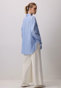 Donna con capelli biondi raccolti indossa una camicia larga a righe blu e bianche, pantaloni bianchi larghi e scarpe bianche, in piedi contro una parete neutra.