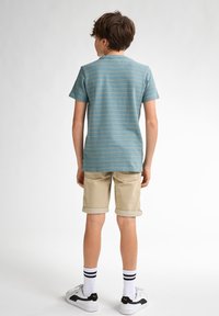 Camiseta a rayas en color verde azulado con mangas cortas, combinada con pantalones cortos beige enrollados. Calcetines blancos con rayas negras y zapatillas con detalles en negro.