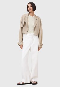 Giacca in suede beige tagliata sopra una maglietta chiara, abbinata a pantaloni bianchi a vita alta e gamba larga, e sandali neri con dettagli in argento.
