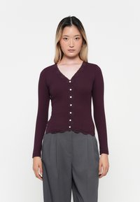 Gilet bordeaux à manches longues avec un décolleté en V bordé de dentelle, doté d'un tissu texturé et de boutons en perles sur le devant, assorti à un pantalon gris.