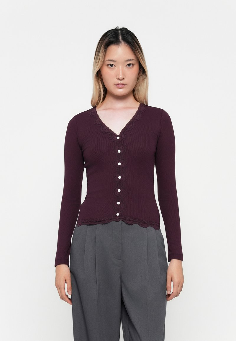 Gilet bordeaux à manches longues avec un décolleté en V bordé de dentelle, doté d'un tissu texturé et de boutons en perles sur le devant, assorti à un pantalon gris.