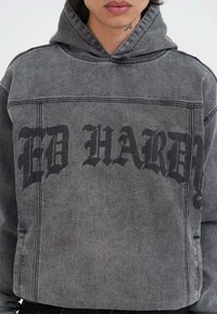 Felpa cappuccio grigio carbone con testo nero stampato in evidenza "ED HARDY." Presenta una vestibilità rilassata e una texture classica simile al denim.