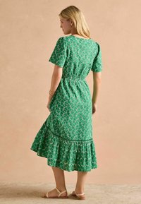 Zelené šaty so vzorom, krátkymi rukávmi, elastickým pásom a volánkovým lemom. Tkanina má hladkú textúru a dekoratívne vzory.