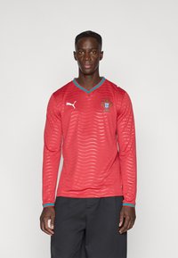 Κόκκινο μακρυμάνικο φανέλα με κυματιστό σχέδιο, αντίθετο τουρκουάζ γιακά και μανσέτες, διαθέτει λογότυπο Puma και έμβλημα της Πορτογαλίας στο στήθος.