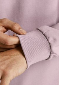 Ljuslila sweatshirtärm med ribbstickad textur och elastisk finish. Hand som greppar tyget visar mjukheten och flexibiliteten hos materialet.