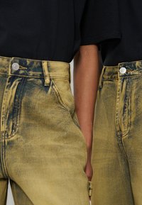 Δύο άνθρωποι με ίδια μαύρα πουκάμισα και κίτρινα τζιν με όξινη πλύση, στέκονται κοντά με εμφανή τα χέρια τους ανάμεσά τους.