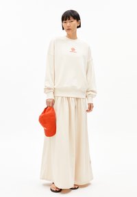 Crème sweatshirt met een rode bloemenlogo, gecombineerd met een luchtige beige rok. Houdt een oranje pet vast. Minimalistisch ontwerp en zachte stof.