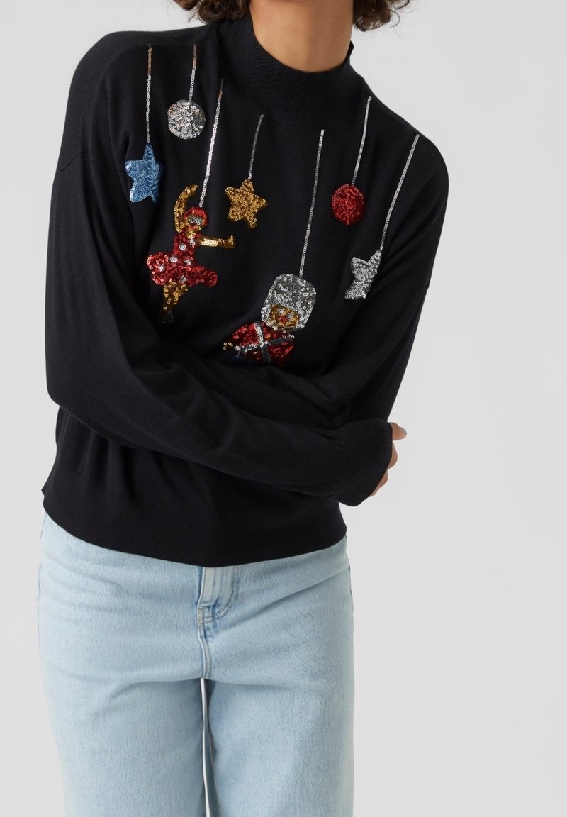 Pull noir avec des ornements suspendus colorés en sequins comprenant des étoiles, une ballerine et un soldat, porté avec un jean bleu clair.