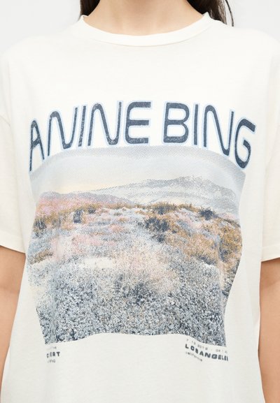 Γυναίκα που φοράει λευκό t-shirt με το κείμενο «ANINE BING» και ένα pixelated τοπίο στην μπροστινή πλευρά.