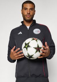 Veste noire zippée avec des accents rouges, arborant un logo. Tenant un ballon de football blanc avec des étoiles noires et des détails colorés.
