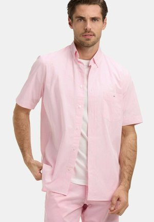 Homme portant une chemise rose clair à manches courtes boutonnée sur un t-shirt blanc et un pantalon rose assorti, debout devant un fond uni.