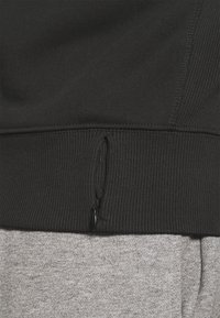 Svart sweatshirt med ribbad nederkant och en funktionell dragkedjeficka. Mjuk grå tygstruktur syns under, med kontrasterande färg.