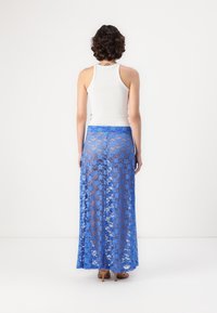 Jupe maxi en dentelle bleue avec un motif floral, texture translucide et taille élastique. Portée par-dessus un débardeur blanc, avec vue de dos visible.