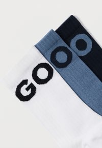 Τρία ζευγάρια κάλτσες: ένα λευκό με μαύρο κείμενο "GOO", ένα ανοιχτό μπλε με σκούρο μπλε γράμμα "O" και ένα σκούρο μπλε. Μαλακή, πλεκτή υφή.