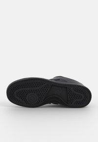 Semelle noire de sneaker avec un motif ondulé, détail circulaire près des orteils et logo New Balance dans la zone de la voûte centrale.