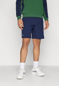 Uomo che indossa pantaloni corti blu navy con il logo Lacoste, felpa verde e blu navy, calze bianche e sneakers Joma bianche, in piedi su un pavimento grigio.