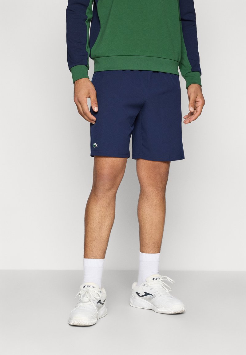 Uomo che indossa pantaloni corti blu navy con il logo Lacoste, felpa verde e blu navy, calze bianche e sneakers Joma bianche, in piedi su un pavimento grigio.