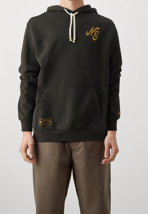 Person iført en sort hættetrøje med hvide snore, goldt "NE" logo på brystet, forlomme og brune bukser, stående foran en ensfarvet baggrund.