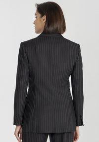 Czarna marynarka w prążki, z wyraźnymi ramionami, slim fit, z zapięciem na jeden guzik i kieszeniami w szwach. Materiał ma gładką teksturę.