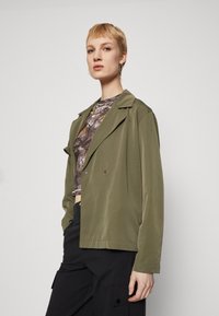 Olive green, lightweight jacket with a relaxed fit, shawl collar, double buttons, and side pockets, worn over a patterned cropped top.