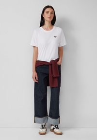 T-shirt en coton blanc avec un patch en forme de cœur, associé à un pantalon en denim bleu large avec un ourlet retroussé et un pull bordeaux noué à la taille.